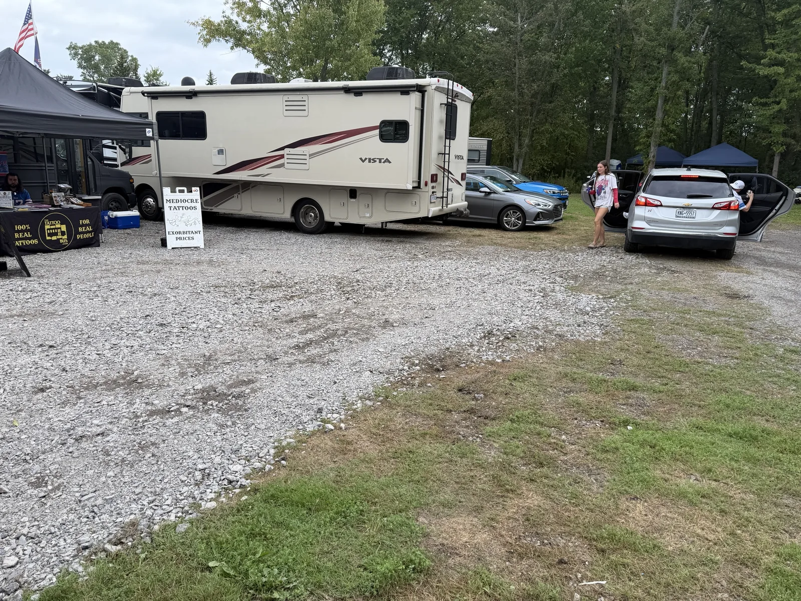 The gravel and grass parking lot with an RV, cars and canopies, backing onto a treeline.
